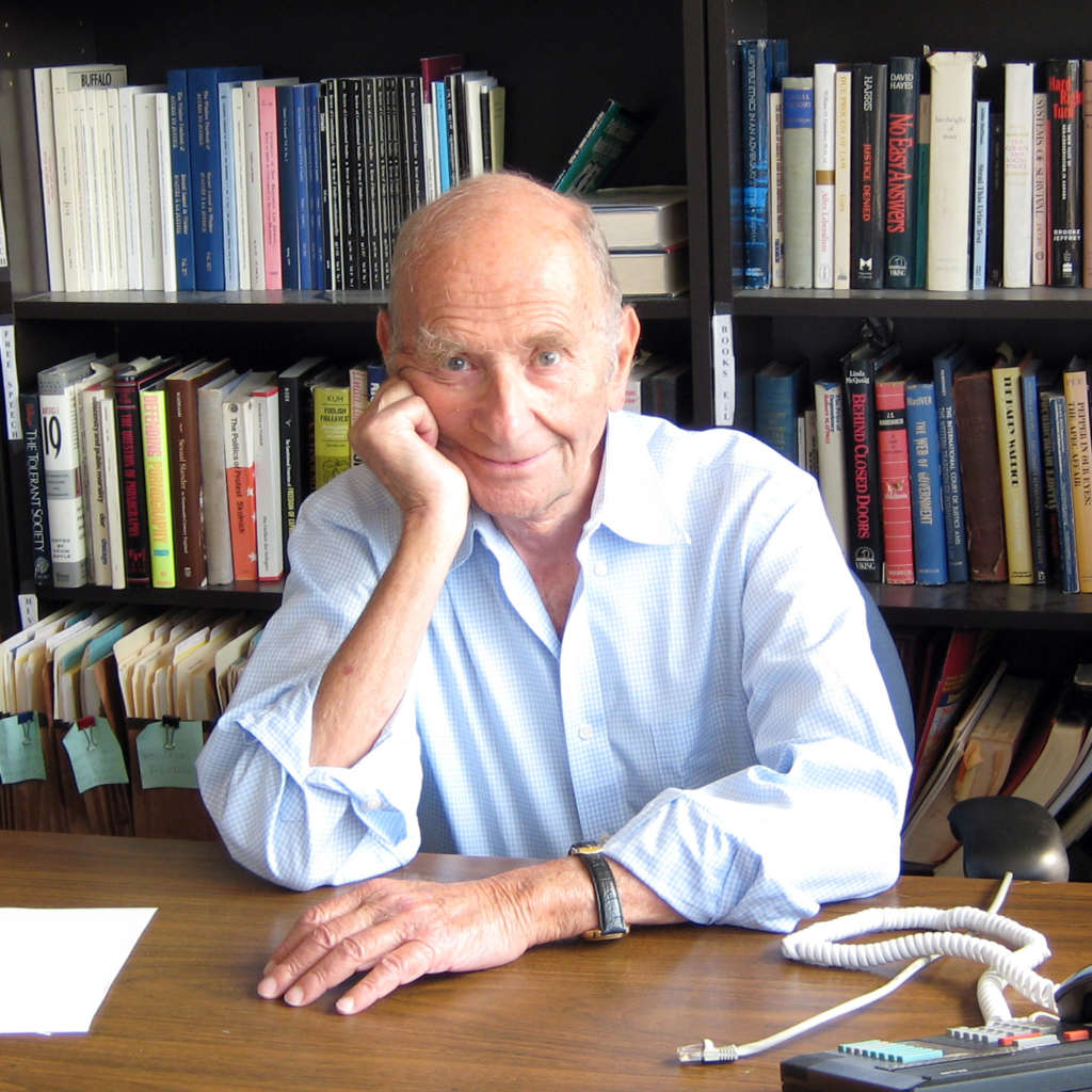 Alan Borovoy sitting at his desk with many books in the background. He is smiling and is leaning his right cheek against his hand.