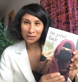 Natasha Bakht holding a copy of her book, In Your Face. She has a medium skin tone and black hair, and she is wearing a white blazer.